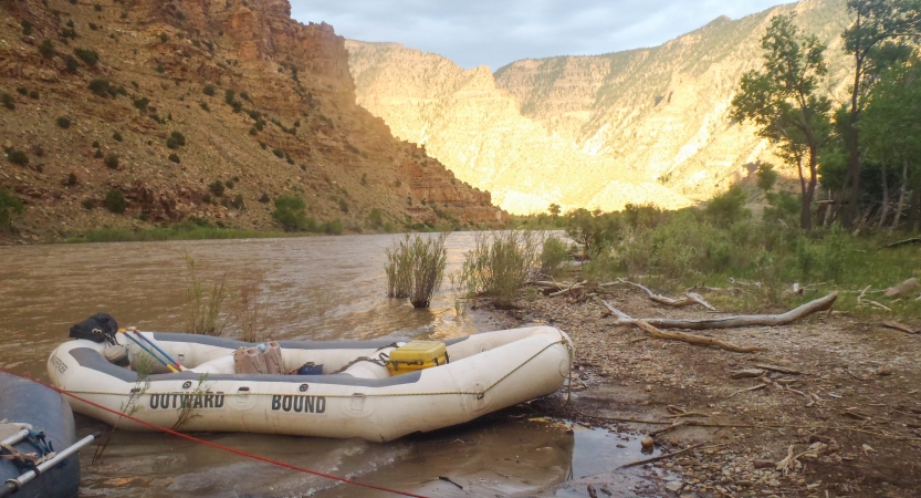 An empty raft rests on the shore of a river. In the background there are tall canyon walls.